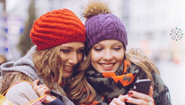 two women wearing knit hats and scarves looking at a phone