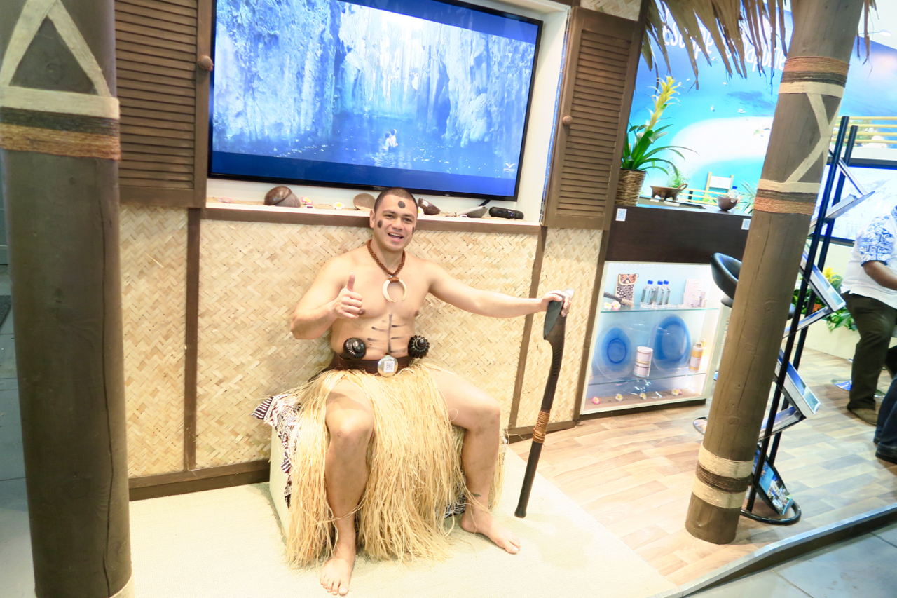 a man sitting on a grass skirt with a large tv in the background