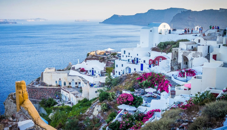 a white buildings on a hill by the ocean with Santorini in the background